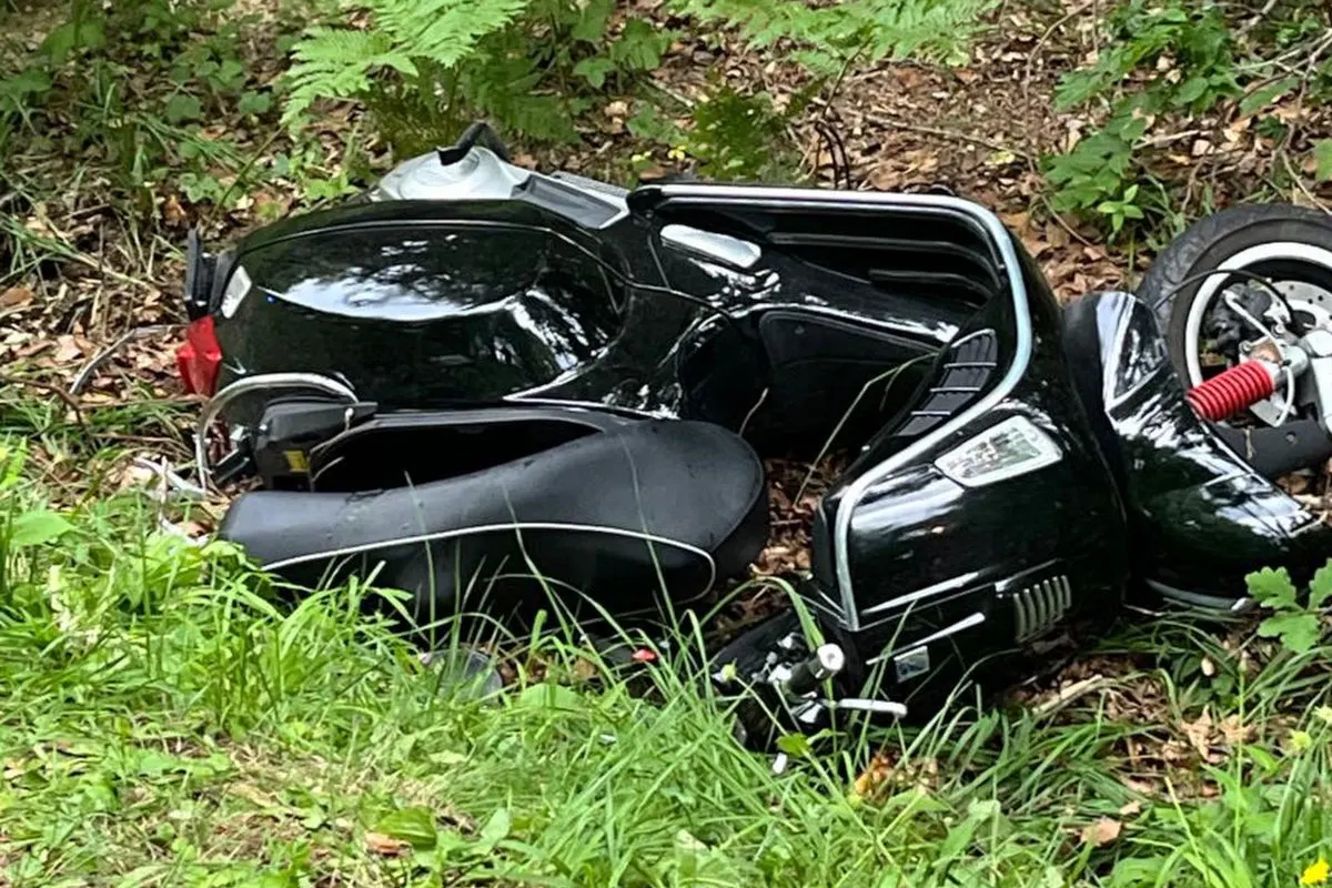Ein gestürzter Motorroller liegt auf der Seite in einem Waldgebiet, umgeben von Gras und Laub.