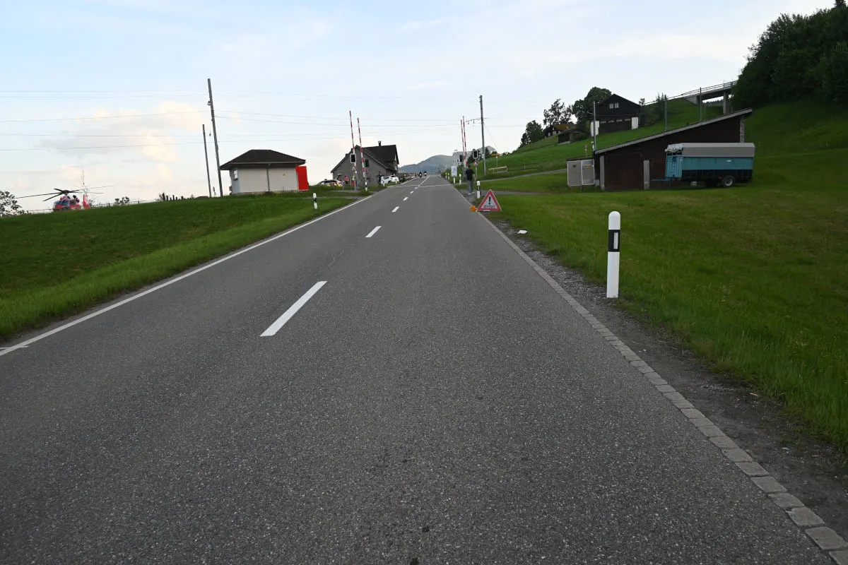 Eine asphaltierte Landstraße führt geradeaus, mit einem Rettungshubschrauber, der auf einem Feld links neben der Straße steht. Weiter im Hintergrund sind Bauernhäuser erkennbar.