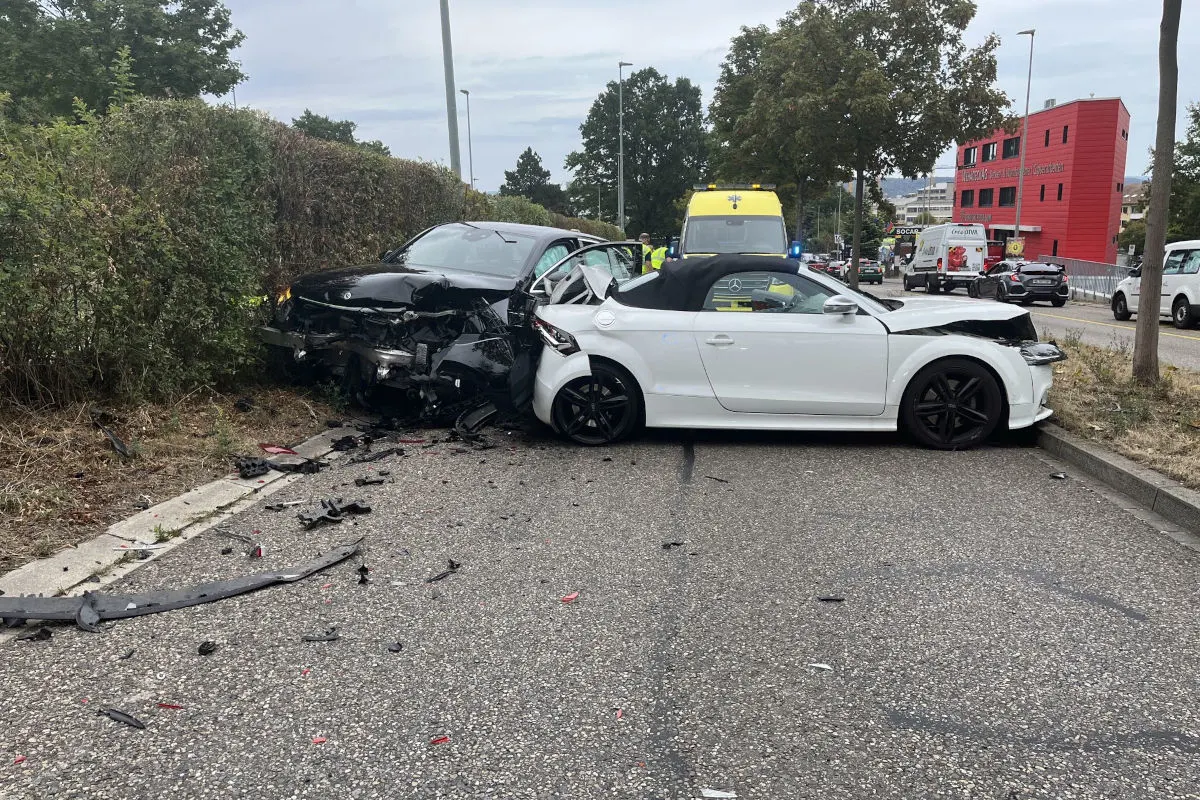 Ein schwarzes und ein weißes Auto sind frontal kollidiert und stehen verbeult auf einer Straße. Ein Krankenwagen ist im Hintergrund zu sehen.