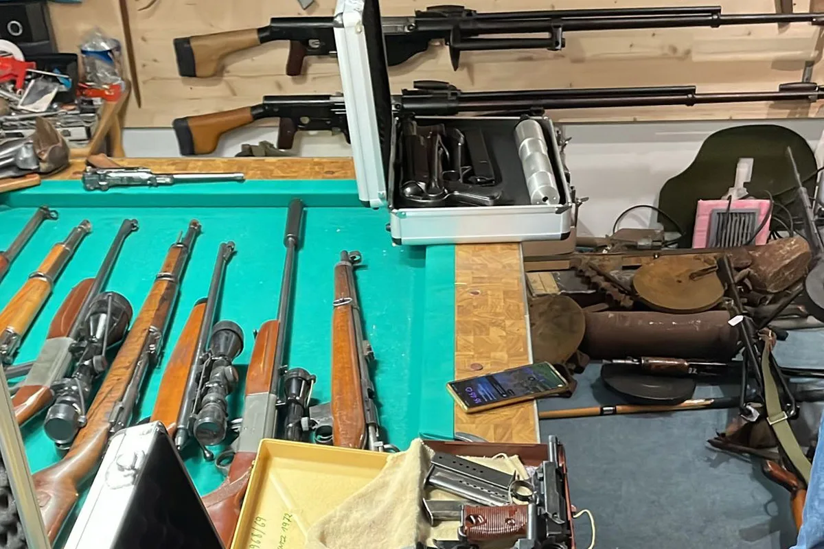 Ein Tisch zeigt zahlreiche Gewehre und Pistolen. Im Hintergrund sind weitere Waffen und Ausrüstungsgegenstände sichtbar.