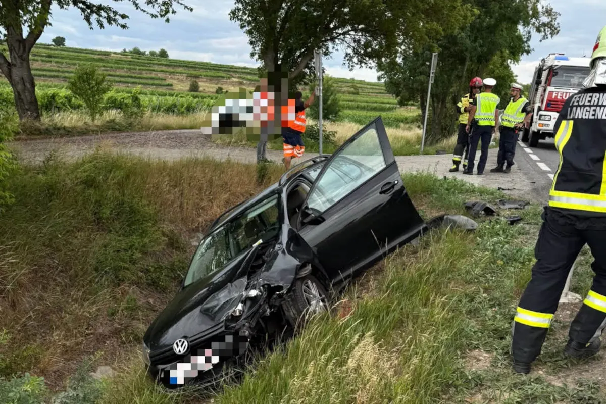 Ein schwarzes Auto liegt beschädigt in einem Graben. Feuerwehrleute und ein Polizeibeamter stehen in der Nähe der Straße.