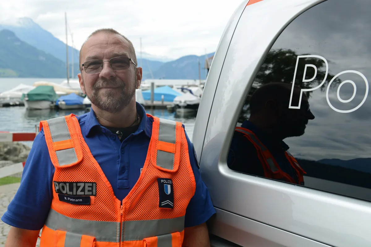 Ein Polizist steht in oranger Weste neben einem silbernen Polizeifahrzeug. Im Hintergrund sind Boote und Berge zu sehen.