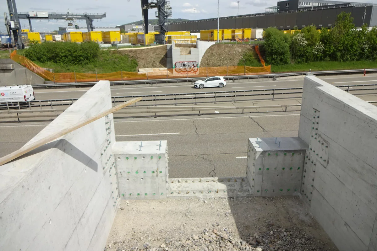 Das Bild zeigt eine im Bau befindliche Autobahnbrücke über einer mehrspurigen Straße. Im Hintergrund sind Industriegebäude und Container zu sehen.