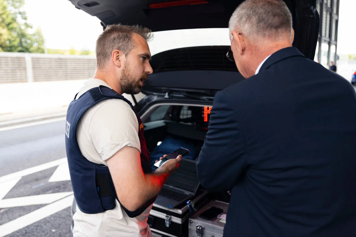 Ein Polizist mit Handy spricht mit einer Person im Anzug am offenen Kofferraum eines Fahrzeugs auf der Autobahn. Im Hintergrund sind Verkehrsschilder zu sehen.