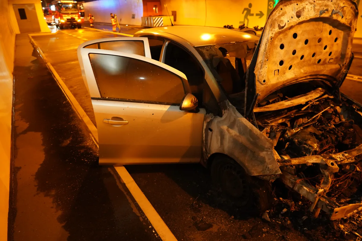 Ein ausgebranntes Auto sieht man in einem Tunnel, das die Feuerwehr umringt. Die Motorhaube und Türe stehen offen.