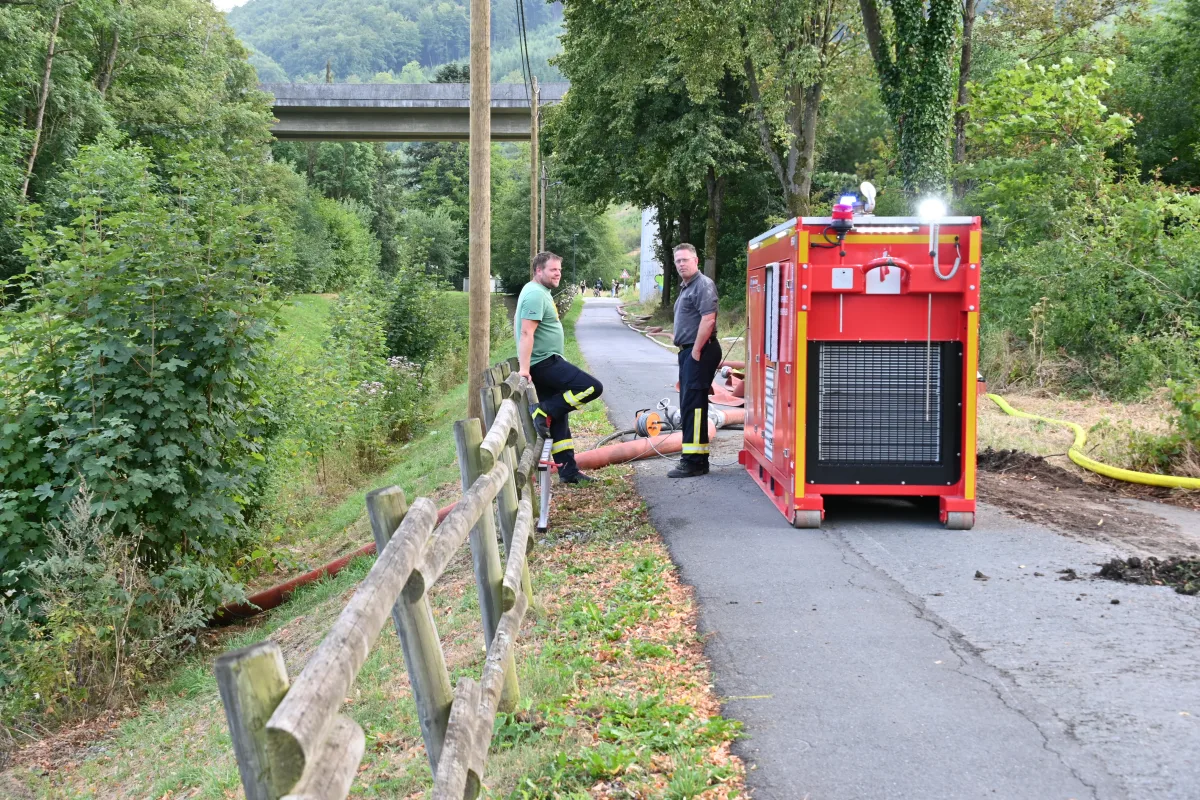 Zwei Männer in Feuerwehrkleidung stehen neben einem roten Gerät auf einem Weg. Im Hintergrund ist grüne Vegetation sichtbar.