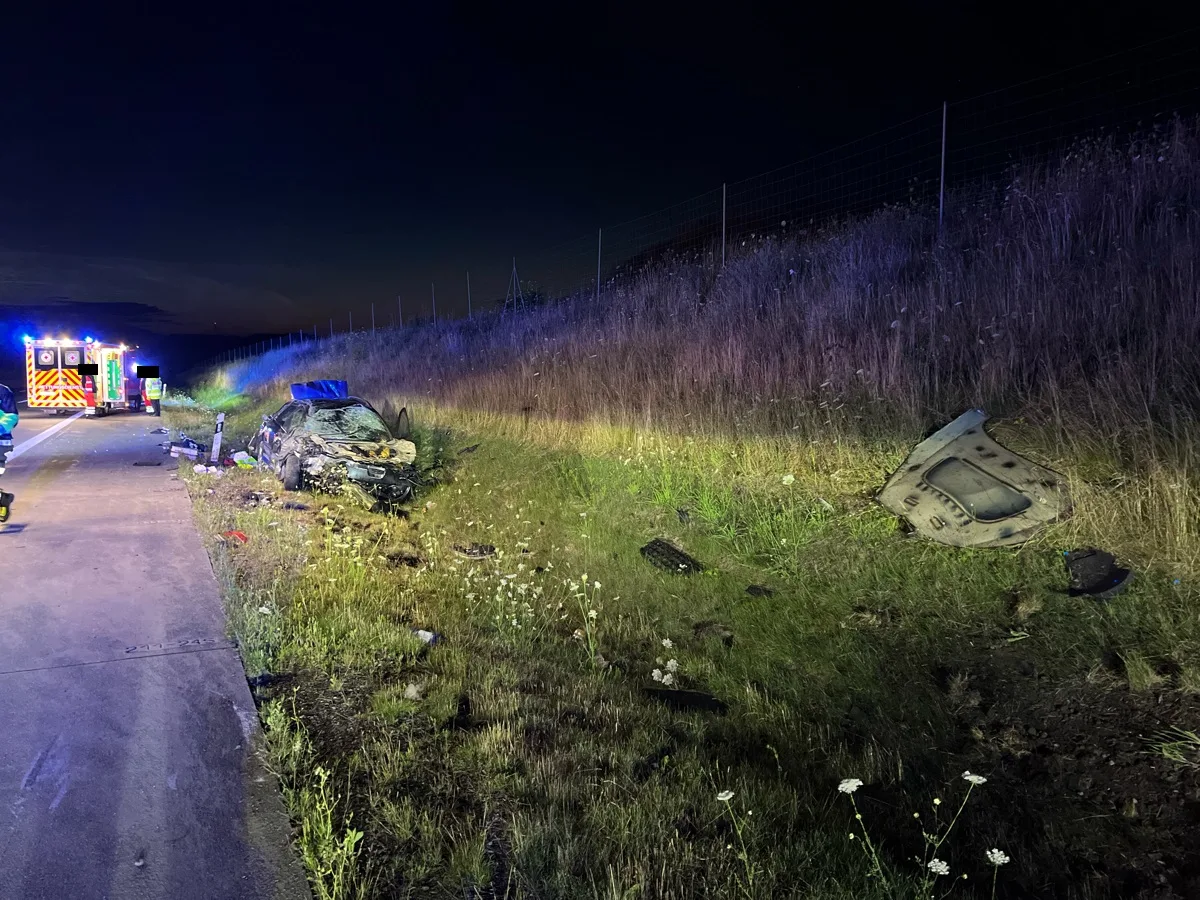 Ein schwer beschädigtes Fahrzeug liegt neben einer Autobahnböschung bei Nacht, während ein Einsatzfahrzeug mit blinkenden Lichtern im Hintergrund sichtbar ist.