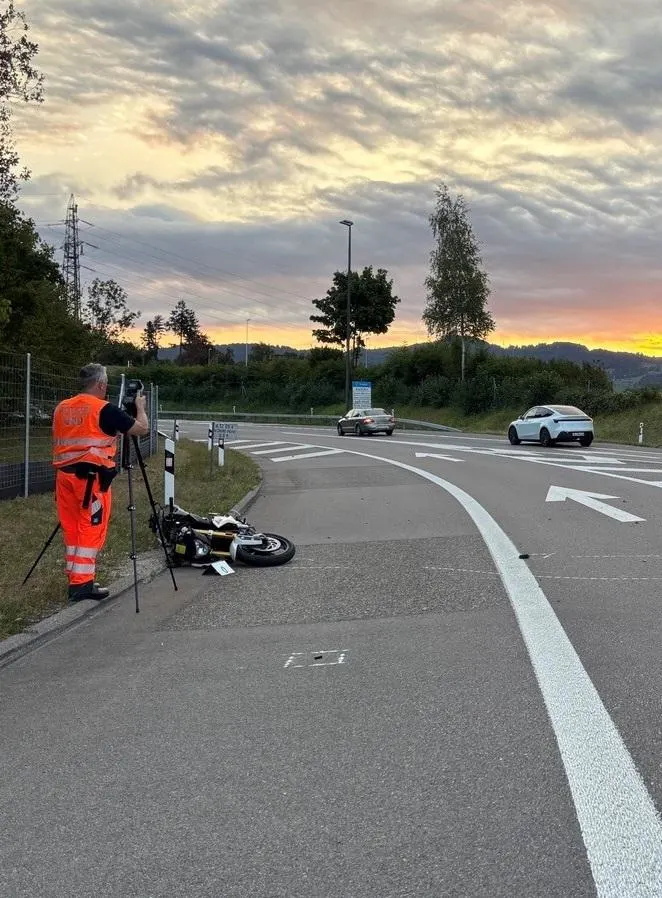 Ein Ermittler in Warnweste untersucht eine Unfallstelle mit einem umgestürzten Motorrad auf einer Landstraße. Autos passieren im Hintergrund, während die Sonne aufgeht.