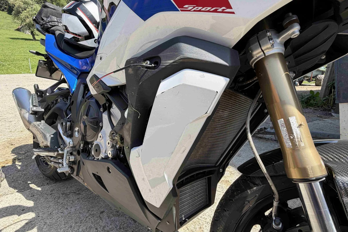 Ein beschädigtes Motorrad mit sichtbaren Kratzern und Dellen an der Verkleidung steht auf einem asphaltierten Untergrund. Im Hintergrund ist eine ländliche Umgebung zu erkennen.