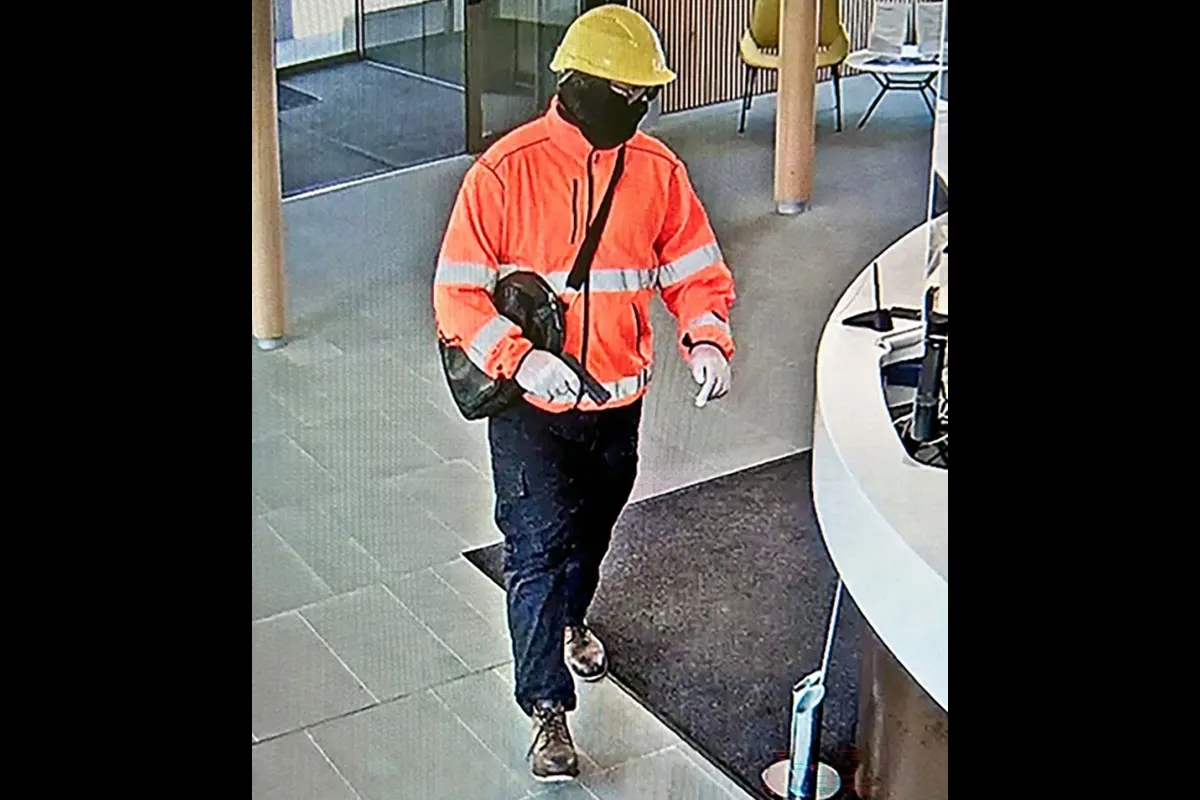 Eine Person in einer orangefarbenen Warnjacke mit reflektierenden Streifen, schwarzer Hose und gelbem Schutzhelm betritt ein Gebäude. Die Person trägt eine schwarze Maske und Handschuhe.