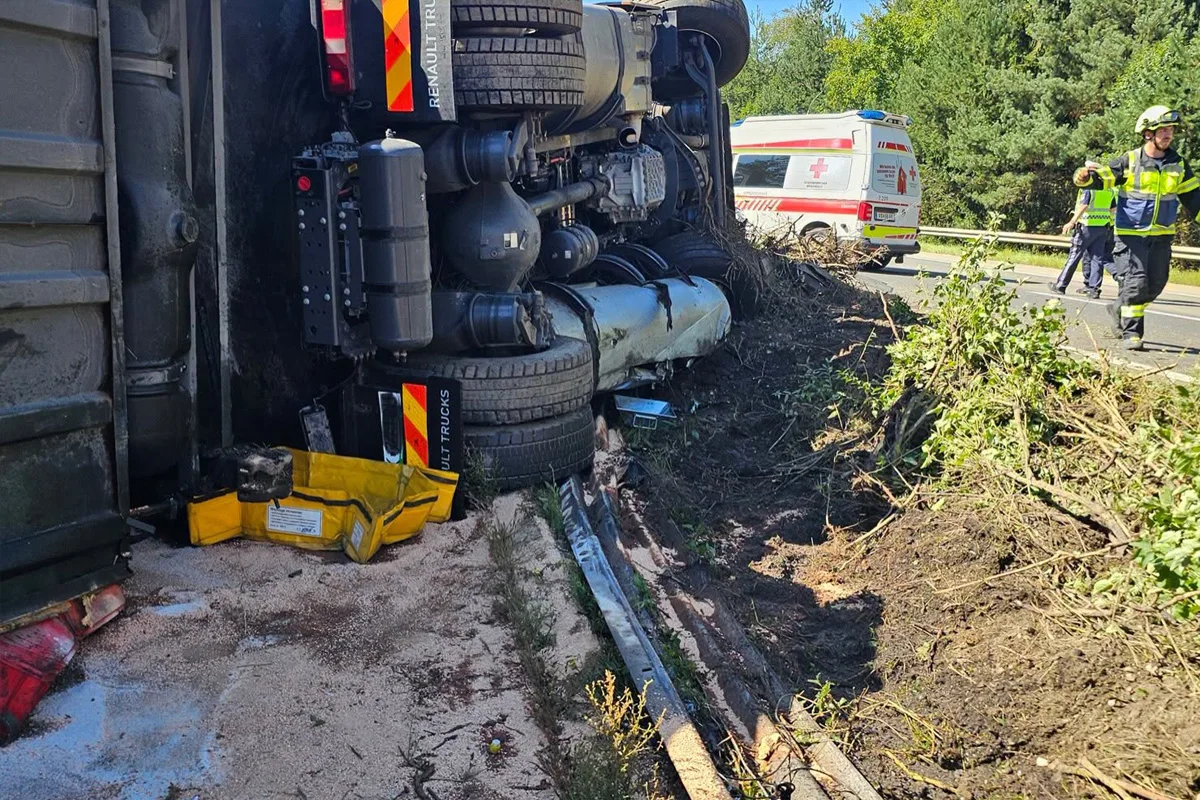 Ein umgestürzter Lastwagen liegt auf der Seite am Rand einer Landstraße. Im Hintergrund steht ein Rettungswagen, während Einsatzkräfte vor Ort sind.