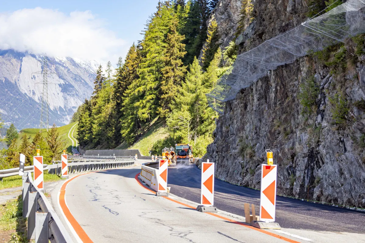 Eine kurvige Bergstraße mit Baustellenfahrzeug und Sicherheitsabsperrungen. Im Hintergrund sind Bäume und ein Felsgitter sichtbar.