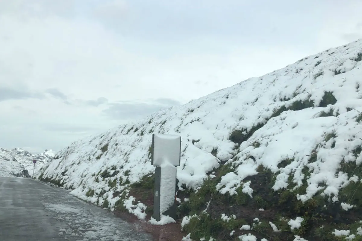 Das Bild zeigt eine verschneite Bergstraße mit einem kaum sichtbaren Verkehrsschild. Der Boden und der Hang sind von Schnee bedeckt.