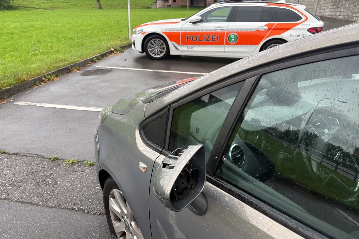 Ein graues Auto mit einem beschädigten Außenspiegel steht auf einem Parkplatz. Im Hintergrund parkt ein Fahrzeug der Polizei mit orange-weißer Markierung.