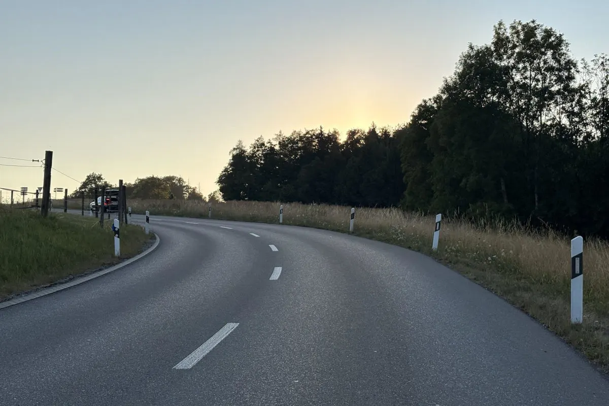 Eine kurvige Landstraße bei Sonnenuntergang, mit einem Fahrzeug am linken Straßenrand. Die Umgebung ist ländlich mit Bäumen am rechten Rand.