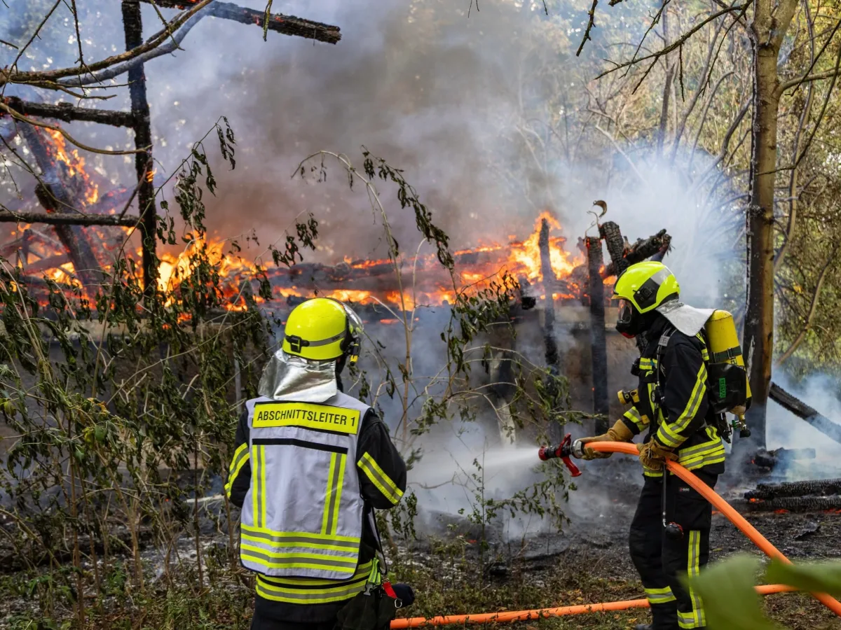 Zwei Feuerwehrleute, erkennbar an ihren Helmen und Schutzanzügen, löschen mit einem Schlauch ein Gebäude im Wald, das in Flammen steht.