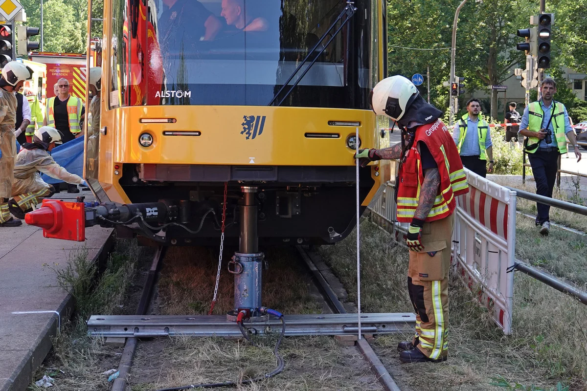 Ein Feuerwehrmann in Schutzkleidung misst den Abstand vor einer gelben Straßenbahn, während weitere Einsatzkräfte anwesend sind.