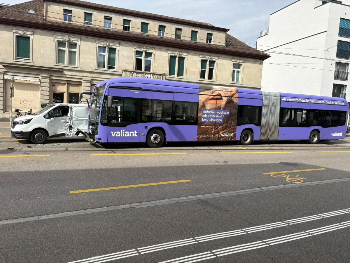 Ein violetter Linienbus ist frontal mit einem weißen Lieferwagen auf einer städtischen Straße zusammengestoßen. Der Lieferwagen zeigt erhebliche Beschädigungen auf der linken Seite.