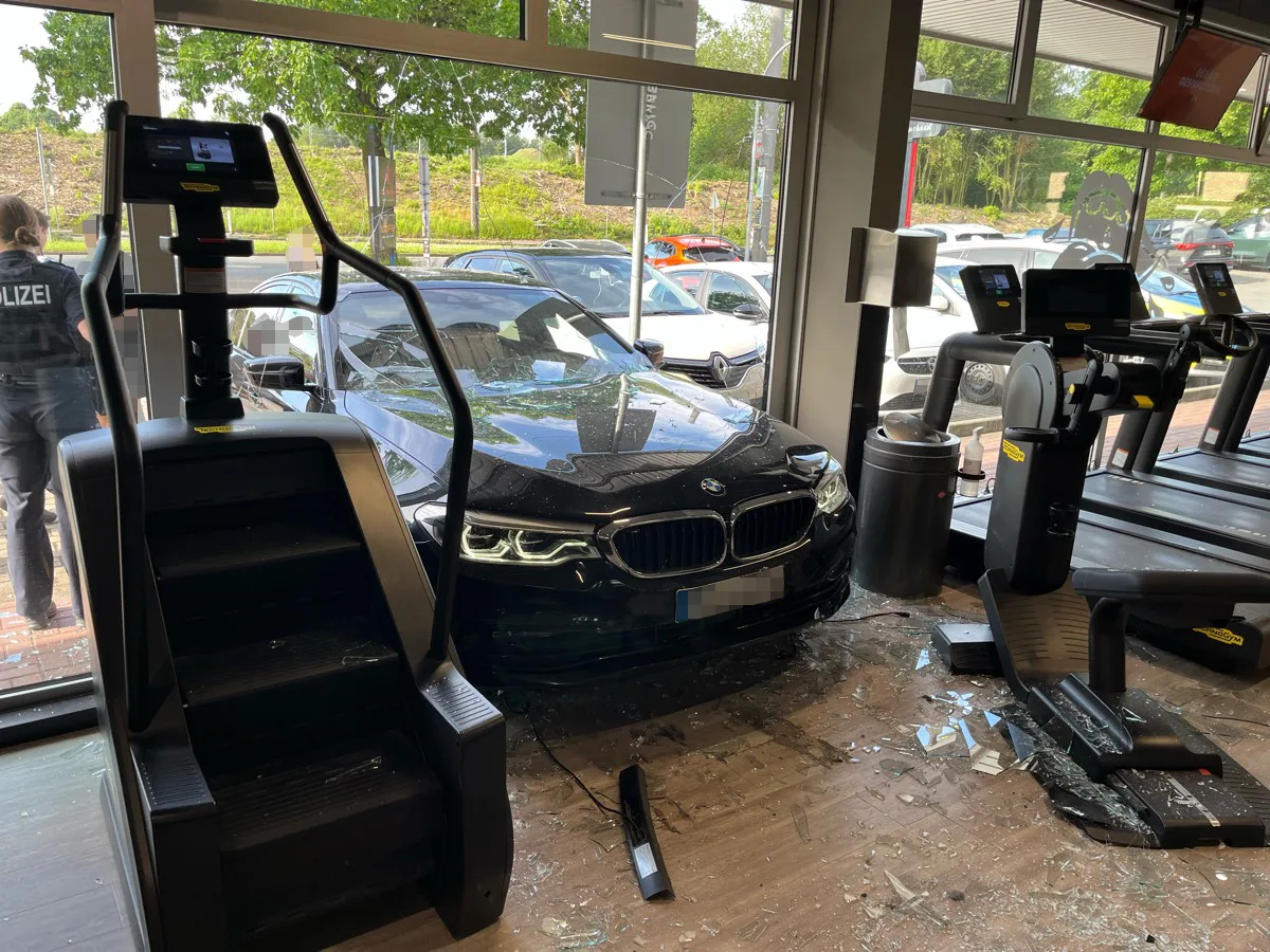 Ein schwarzer BMW ist in ein Fitnessstudio gefahren und hat die Glasfront durchbrochen. Ein Polizeibeamter ist im Hintergrund sichtbar.
