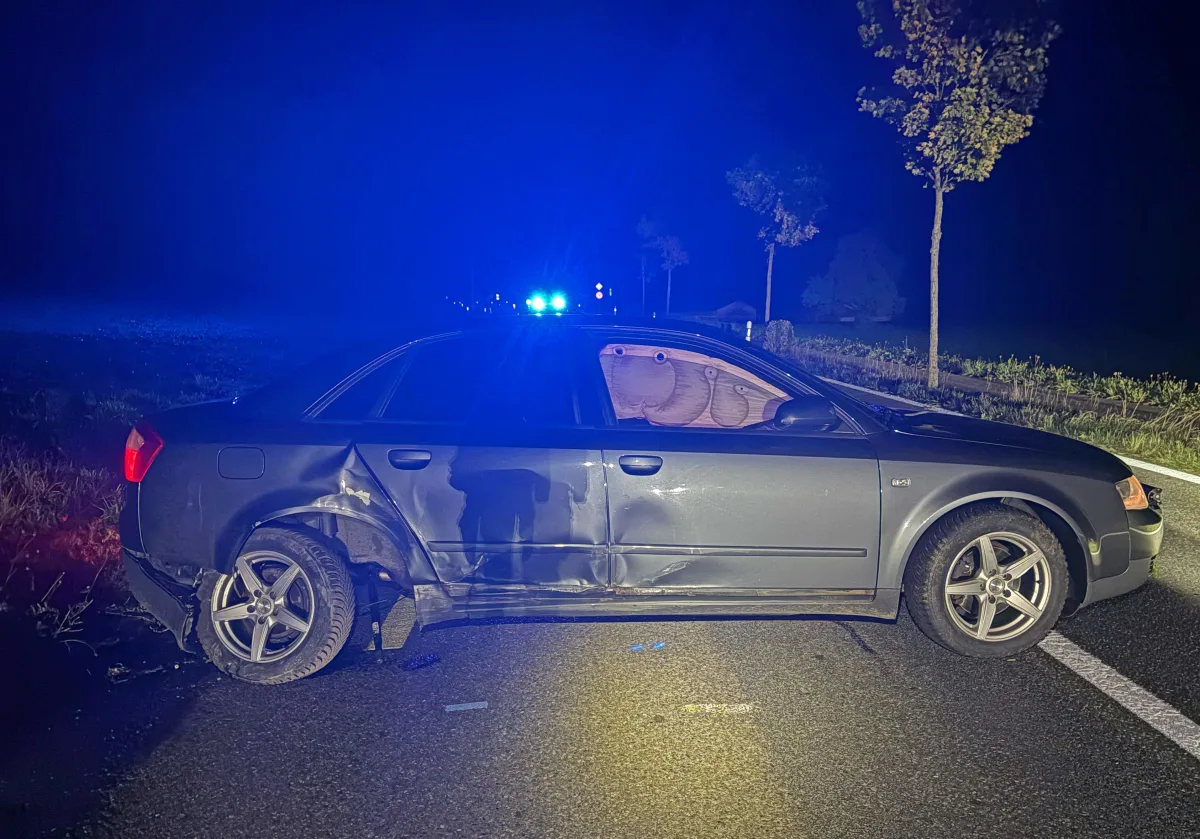Ein parkendes Auto mit sichtbaren Beschädigungen an der Seite steht auf einer Straße bei Nacht. Im Hintergrund leuchtet blaues Licht.