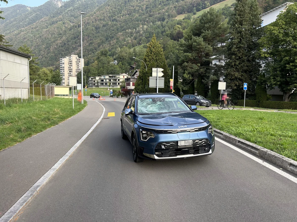 Ein blaues Auto mit zerschlagener Windschutzscheibe steht auf einer Straße mit Berglandschaft im Hintergrund. Es befindet sich in einer ländlichen Umgebung mit weiteren Fahrzeugen und Radfahrern im Hintergrund.