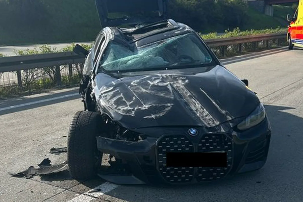 Ein stark beschädigtes schwarzes Auto steht auf dem Seitenstreifen einer Autobahn. Die Frontscheibe ist zerstört und der Wagen hat sichtbare Schäden an der Karosserie.