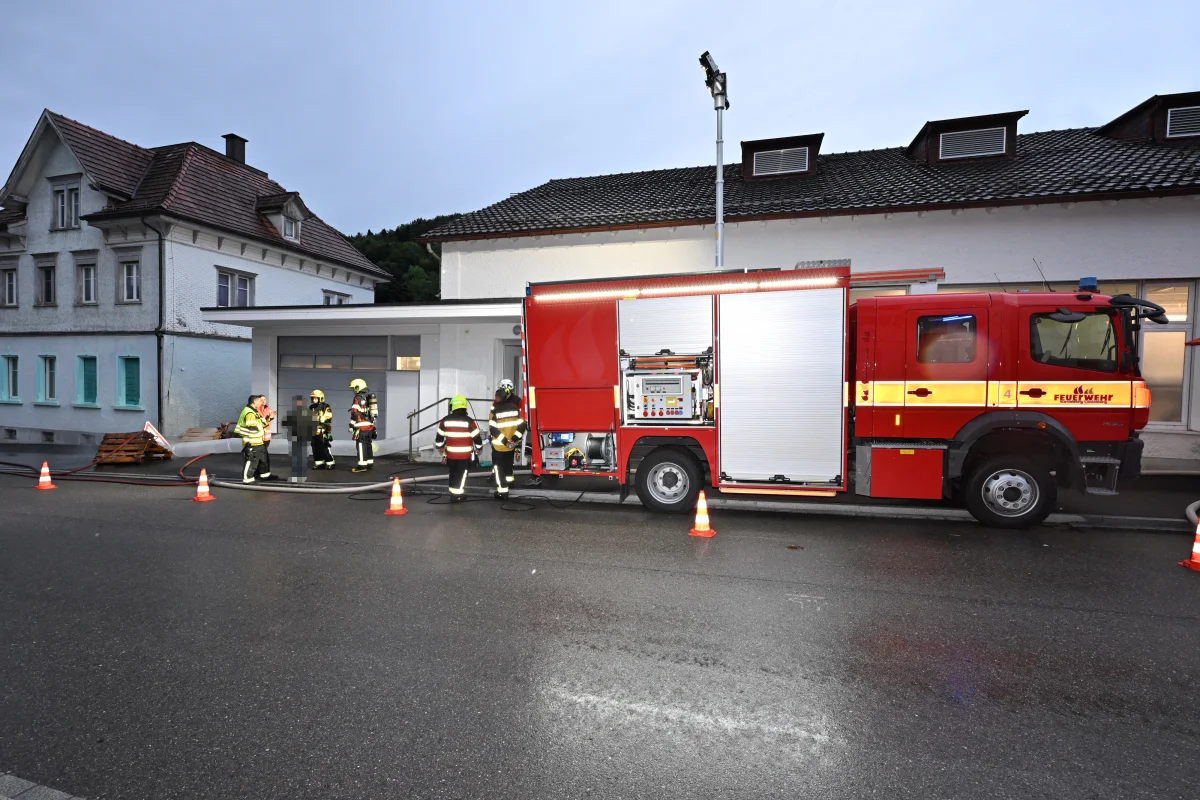 Ein rotes Feuerwehrauto steht vor einem Gebäude, umgeben von Feuerwehrleuten in Schutzkleidung und mehreren orangefarbenen Kegeln.