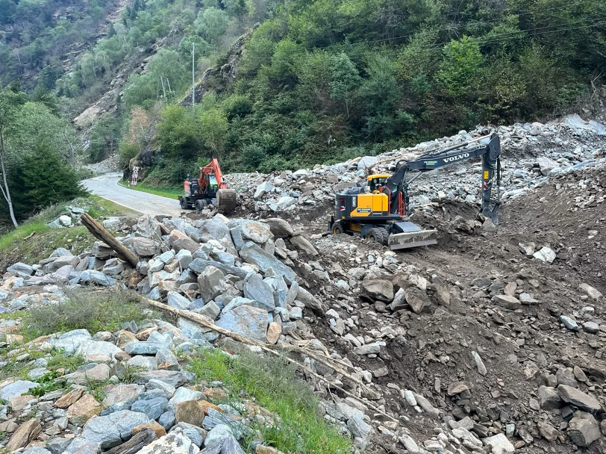 Zwei Bagger arbeiten daran, Geröll und Schutt von einer Bergstraße zu entfernen. Der Bereich ist von Bäumen und felsigem Gelände umgeben.