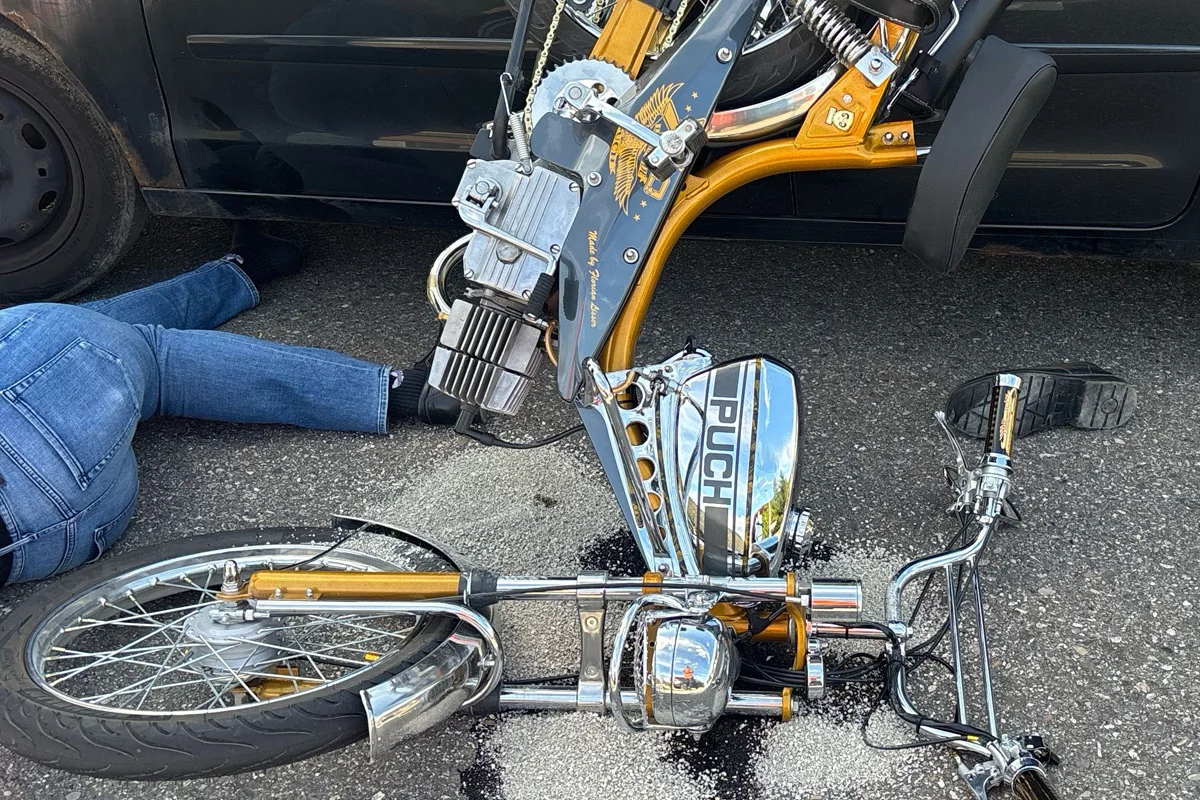 Ein Moped mit der Marke PUCH liegt neben einem Auto auf der Straße. Ein Bein in Jeans ragt unter dem Auto hervor.