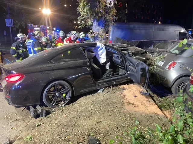 Ein schwarzer BMW ist bei Nacht in einen Unfall verwickelt, die Tür steht offen, umgeben von Feuerwehrleuten. Ein daneben stehender Kleinwagen zeigt Schäden, während Rettungskräfte vor Ort sind.
