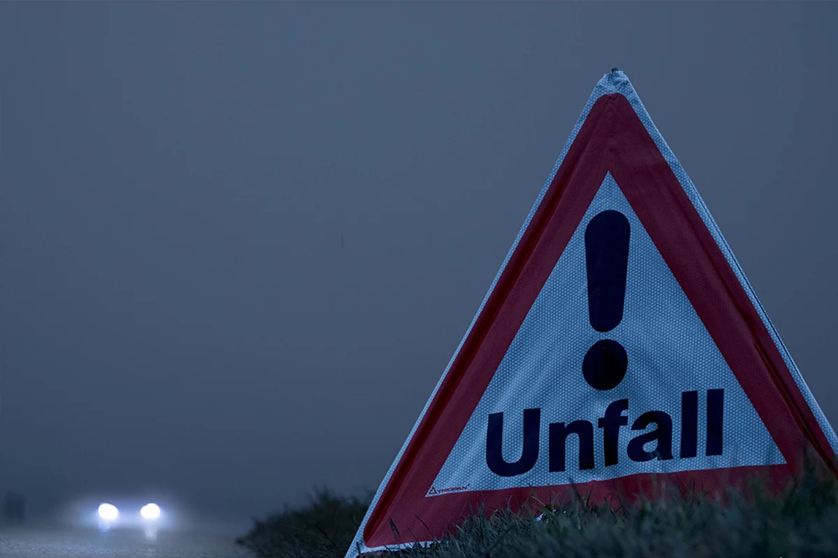 Ein dreieckiges Unfall-Warnschild steht am Straßenrand bei Dunkelheit. Im Hintergrund sind Autoscheinwerfer auf der Straße sichtbar.