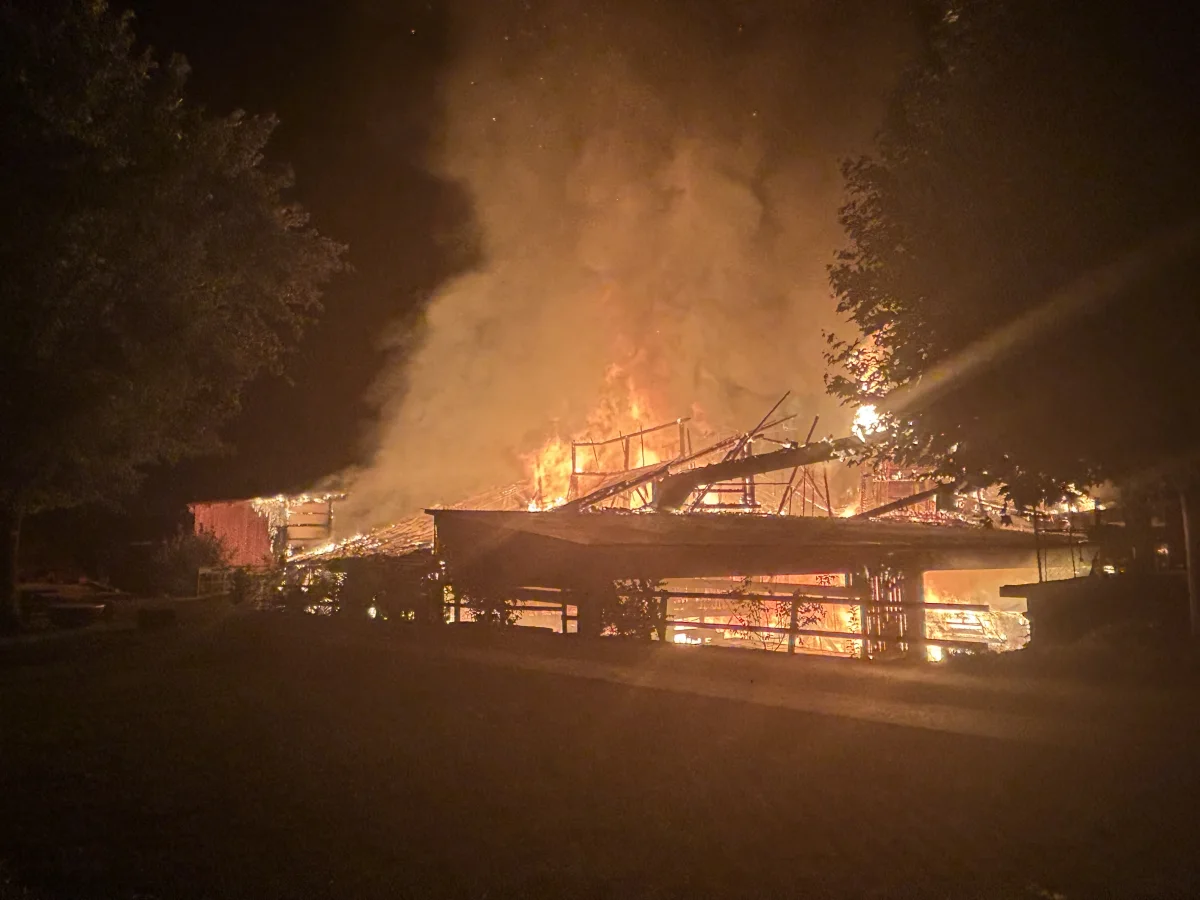 Ein großes Gebäude, vermutlich eine Scheune, steht in Flammen bei Nacht. Flammen und Rauch steigen in den Nachthimmel.