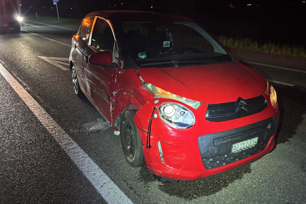Ein rotes Auto mit einem sichtbaren Unfallschaden an der Vorderseite steht auf einer Straße. Der Unfall ereignete sich bei Nacht, die Straße ist wenig beleuchtet.