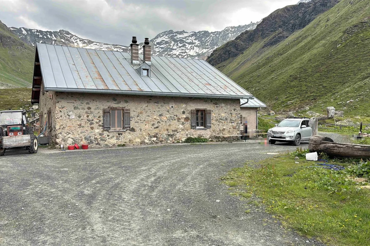 Ein steinernes Haus mit einem Blechdach steht in einer alpinen Umgebung. Ein silbernes Auto ist in der Nähe geparkt. Die Landschaft ist grün, mit Bergen im Hintergrund.
