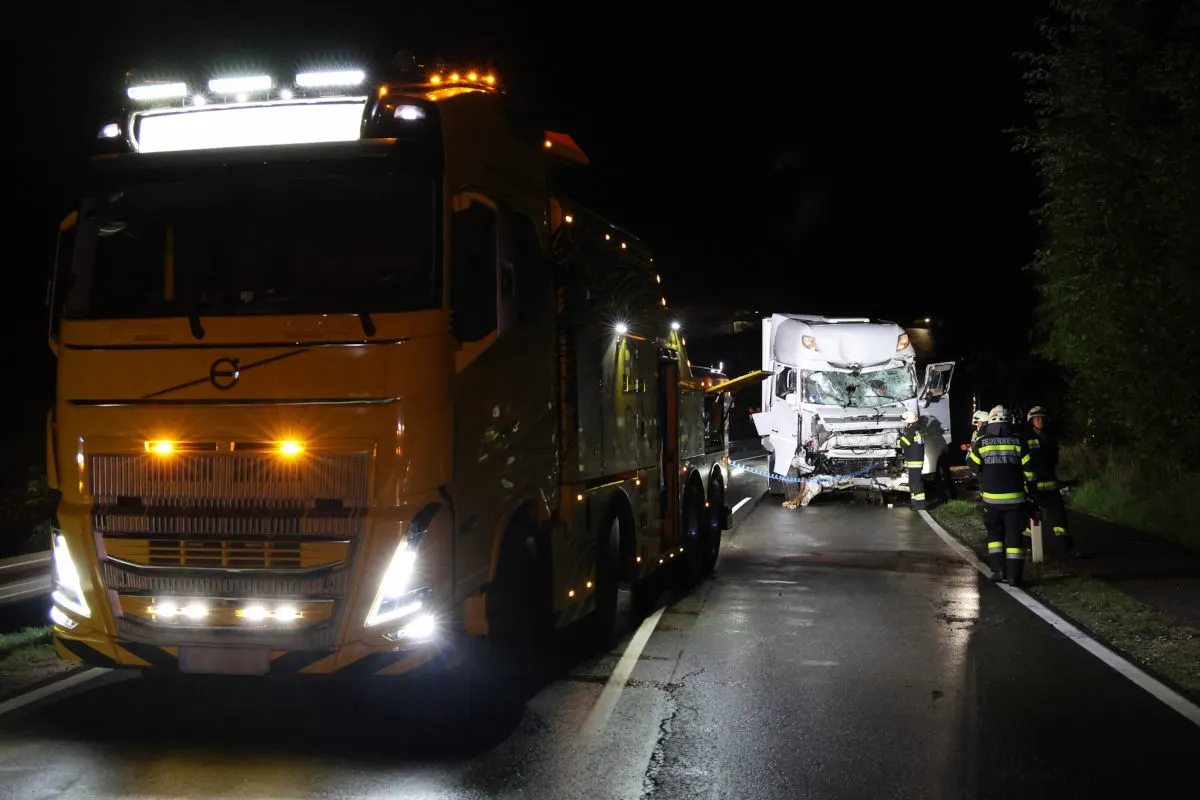 Ein verunfallter Lkw steht neben einem gelben Abschleppwagen auf einer nassen Landstraße bei Nacht. Feuerwehrleute sind ebenfalls anwesend.