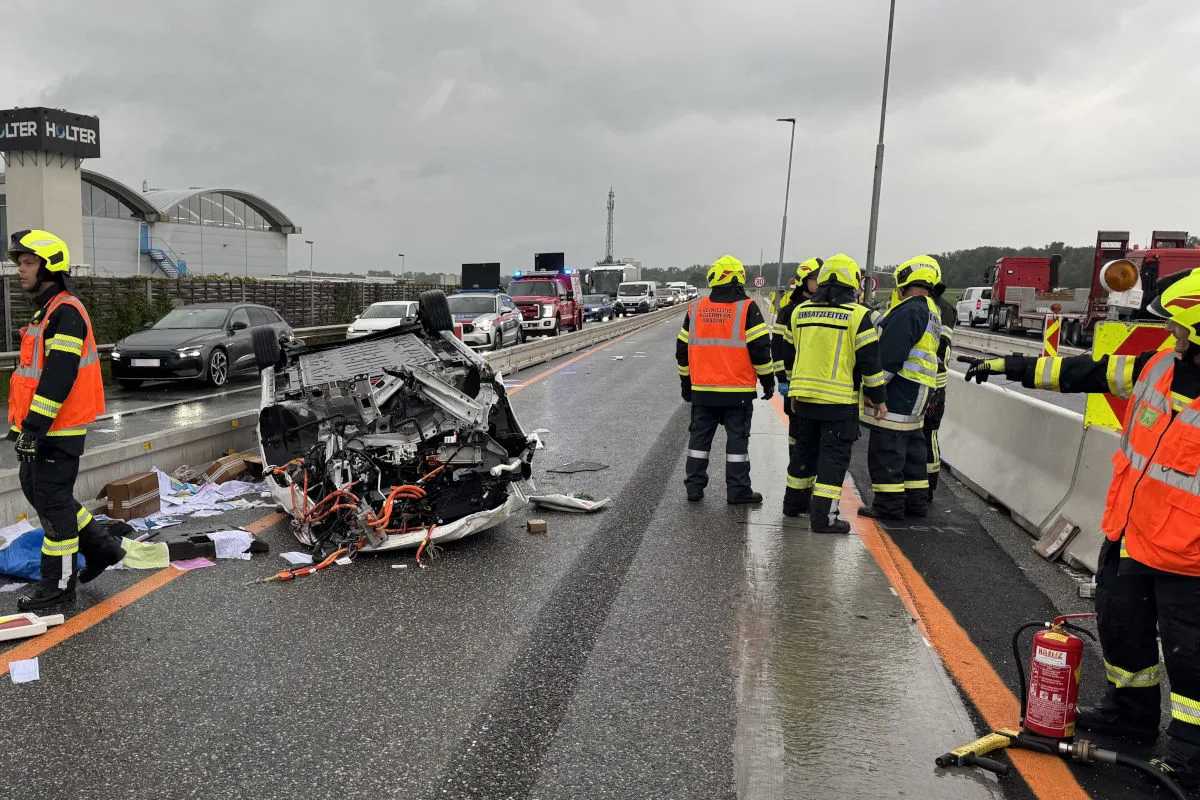 Ein Fahrzeug liegt umgestürzt auf einer Autobahn. Mehrere Feuerwehrleute in Warnwesten stehen um das Fahrzeug herum, während Autos im Stau dahinter stehen.