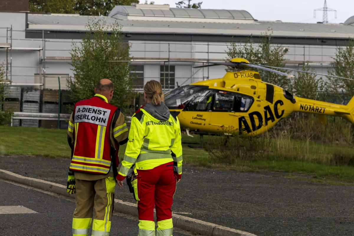 Zwei Personen in Rettungsdienstkleidung stehen auf einer Wiese und blicken zu einem gelben ADAC-Hubschrauber, der gelandet ist. Im Hintergrund ist ein Gebäude zu sehen.