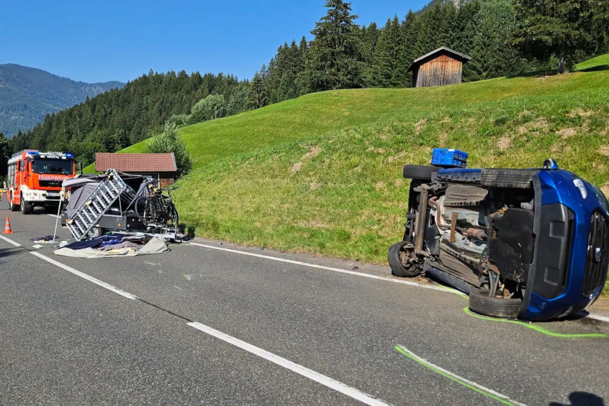 Ein blauer PKW liegt auf der Seite neben einer Landstraße, daneben ein beschädigter Anhänger und ein Feuerwehrfahrzeug.