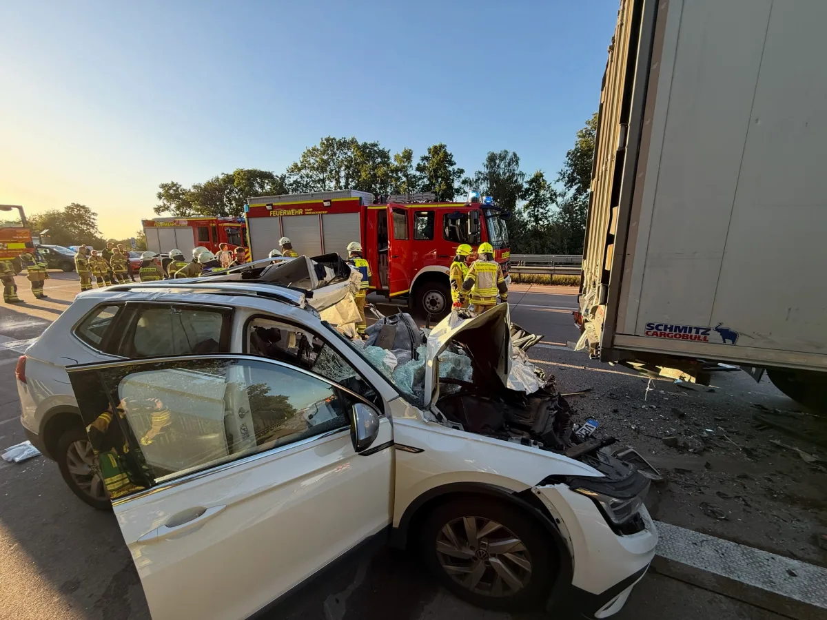 Ein stark beschädigtes weißes Auto ist auf der Autobahn in einen Lastwagen gefahren. Feuerwehrfahrzeuge und Einsatzkräfte sind am Unfallort präsent.