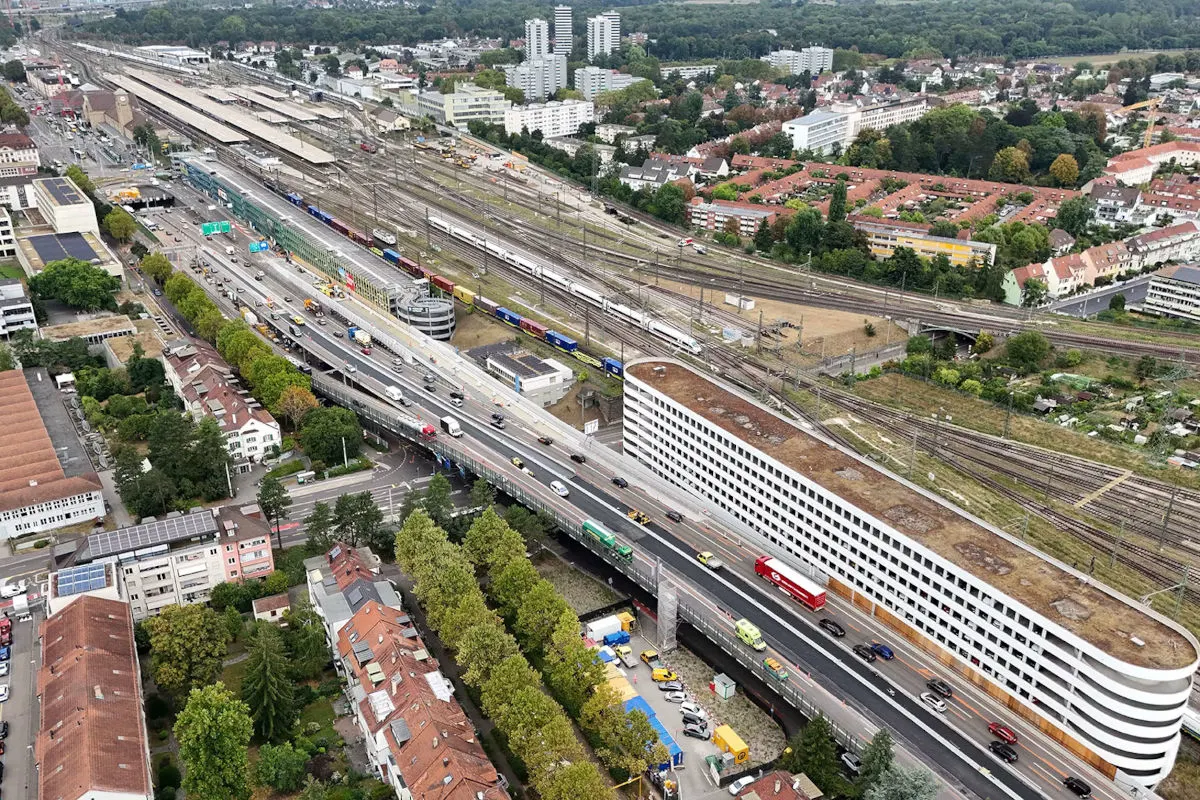 Die Luftaufnahme zeigt eine Stadtlandschaft mit einem großen Bahnhof, mehreren Gleisen und einer Hauptstraße mit Verkehr.