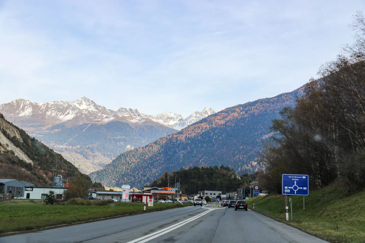 Eine Straße führt durch eine alpine Landschaft mit schneebedeckten Bergen im Hintergrund. Es sind mehrere Fahrzeuge und einige Gebäude entlang der Straße zu sehen.