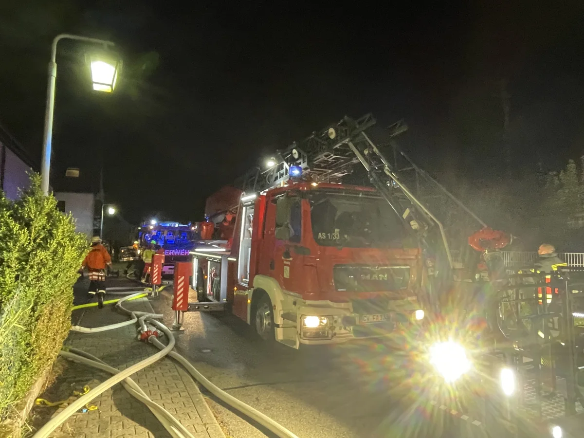 Ein großes rotes Feuerwehrauto mit ausgefahrener Leiter steht nachts auf einer Straße. Feuerwehrleute sind vor Ort.