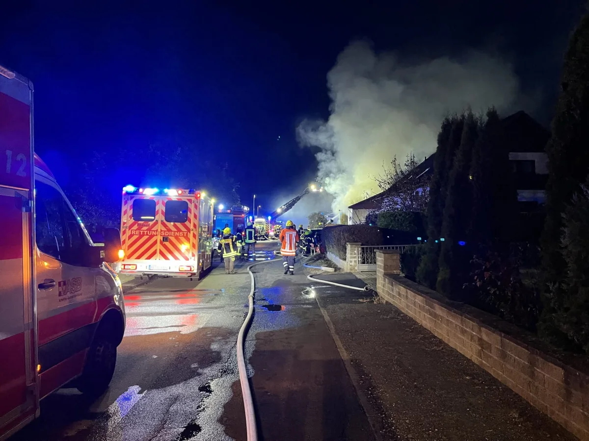 Mehrere Feuerwehr- und Rettungsfahrzeuge sind nachts im Einsatz bei einem Gebäude, aus dem dichter Rauch aufsteigt. Feuerwehrleute in gelber Schutzkleidung arbeiten vor Ort.