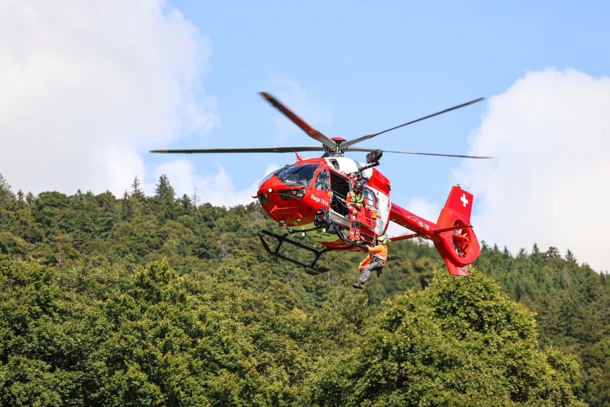 Ein roter Rettungshubschrauber schwebt über einem Wald, zwei Rettungskräfte hängen an einem Seil unter dem Helikopter. Die Szene spielt bei klarem Wetter.