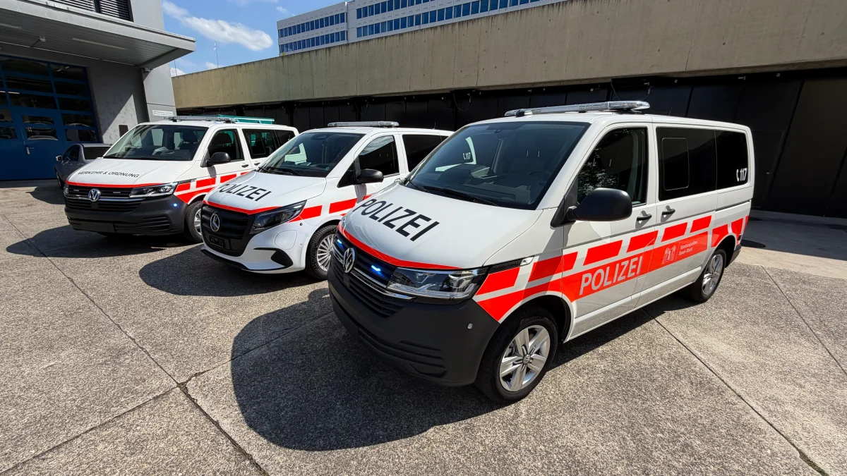 Drei weiße Polizeivans mit der Aufschrift 'POLIZEI' stehen nebeneinander auf einem Parkplatz vor einem Gebäude. Die Fahrzeuge tragen rote Streifen.