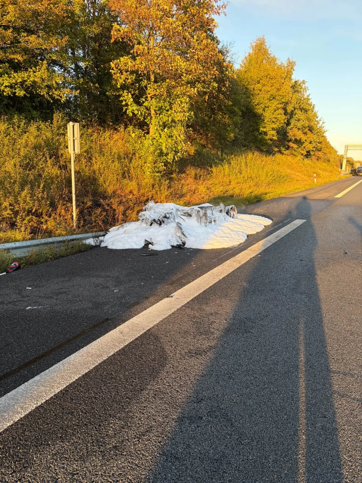 Auf einer Autobahn sind Schaumreste zu sehen, die auf einen Einsatz hindeuten. Im Hintergrund stehen Bäume im warmen Sonnenlicht.