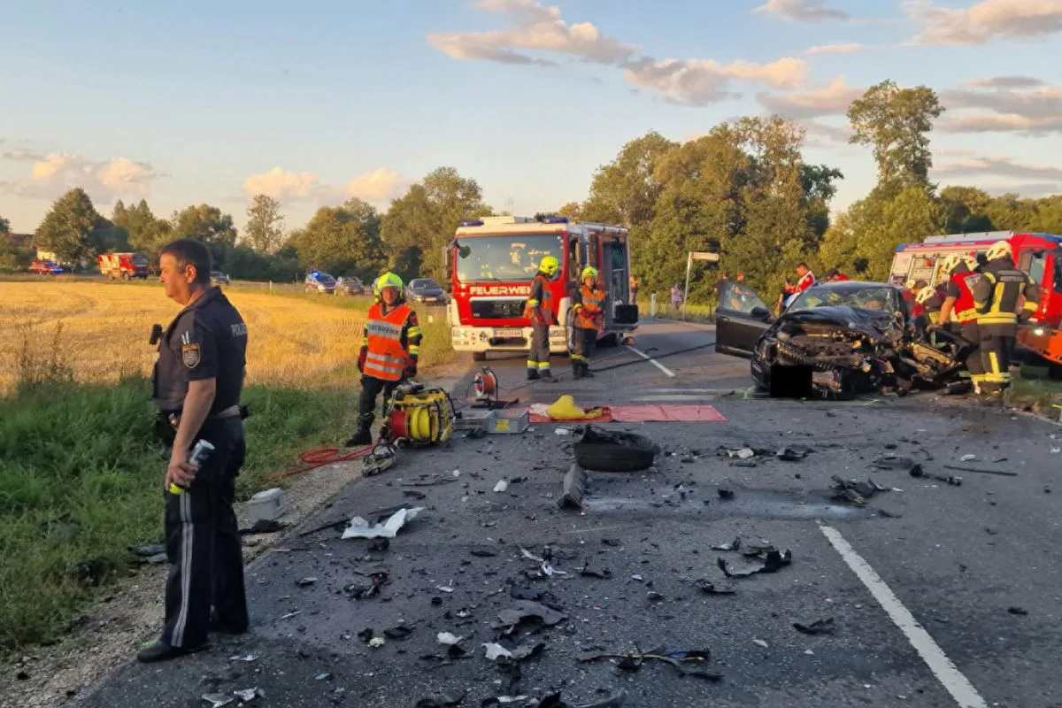 Eine Unfallstelle auf einer Landstraße zeigt ein stark beschädigtes Auto. Feuerwehrleute sind vor Ort, und Trümmer liegen auf der Straße.