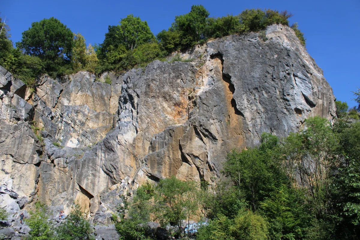 Eine steile Felswand erhebt sich, bedeckt von Bäumen an der Spitze, mit Kletterern an der Wand und dichter Vegetation drumherum.