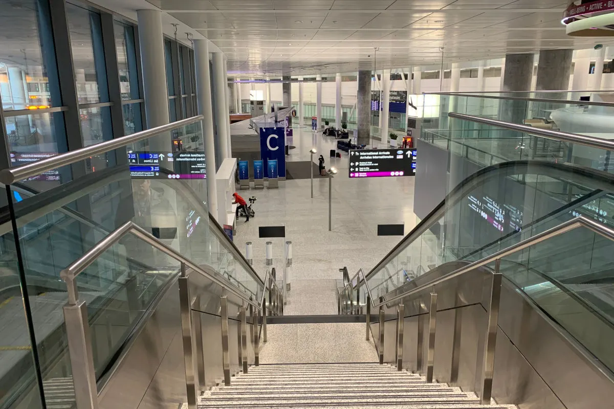 Eine breite Treppe mit Glasgeländer führt in eine großzügige Ankunftshalle eines Flughafens. Anzeigetafeln und Sitzgelegenheiten sind im Hintergrund sichtbar.