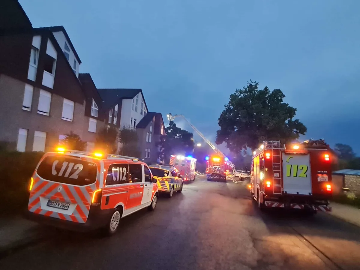 Mehrere Feuerwehrfahrzeuge stehen mit eingeschaltetem Blaulicht vor einer Wohnhäuserreihe. Es ist dämmerig und ein Feuerwehrleiter reicht zum Dach eines Hauses.